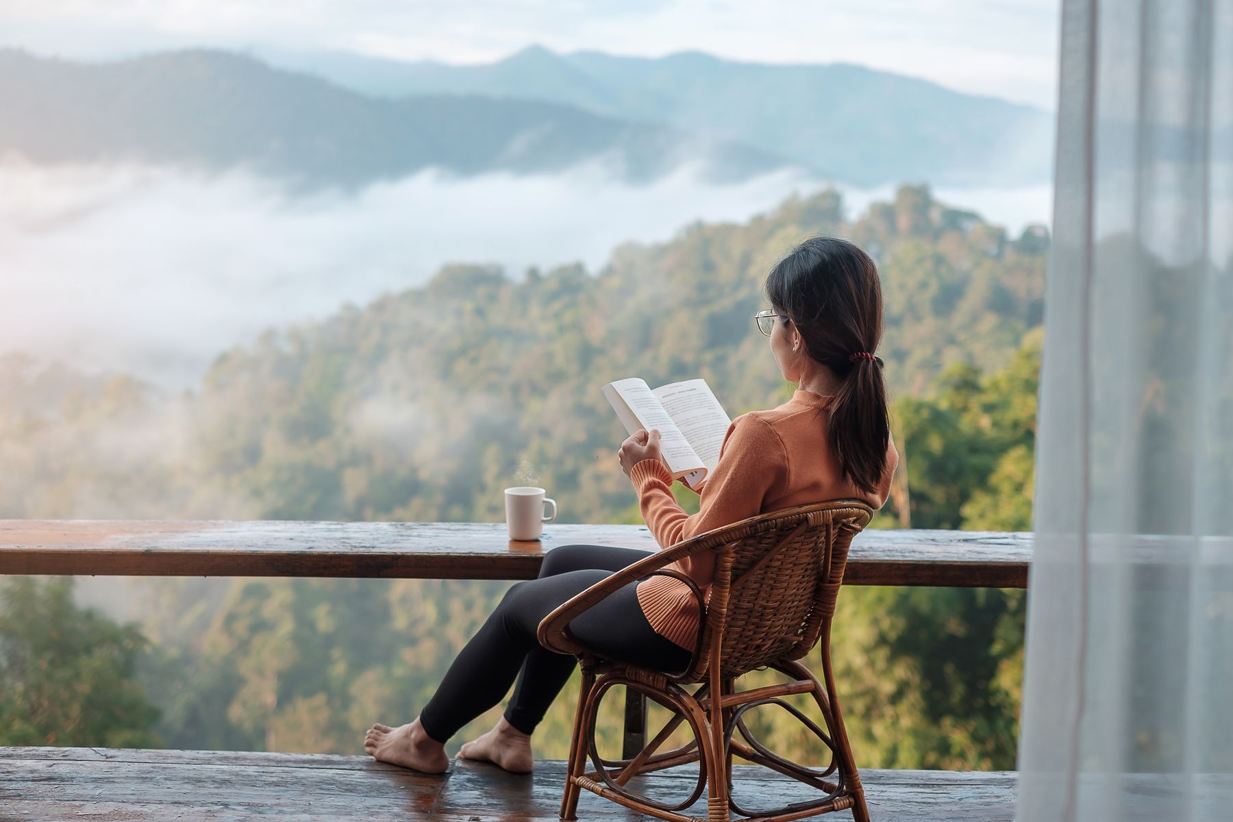 Frau mit Buch auf einer Terrasse mit Blick über bewaldete Berge